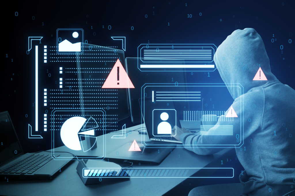 a hooded figure using a laptop with cyber security graphics overlay, symbolizing a cyber attack on a dark blue background